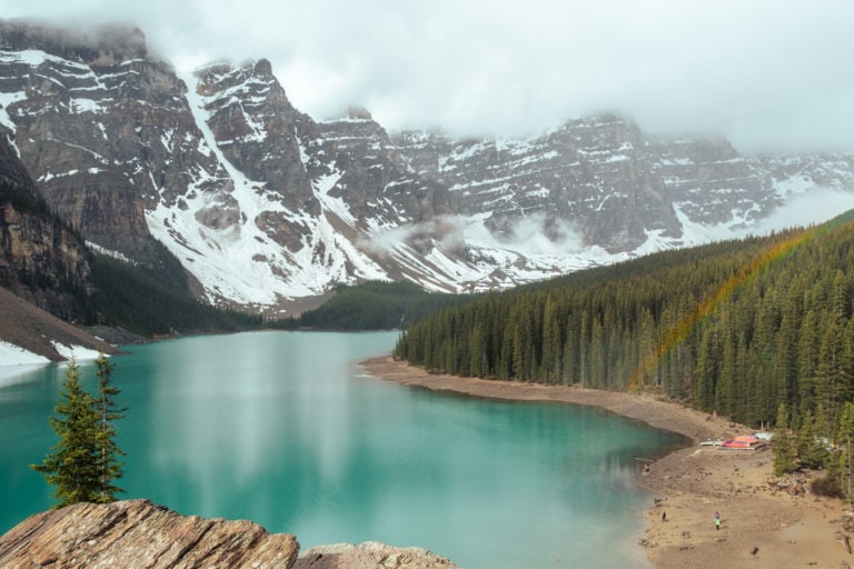 A rainbow over the trees beside the turquoise Moraine Lake.