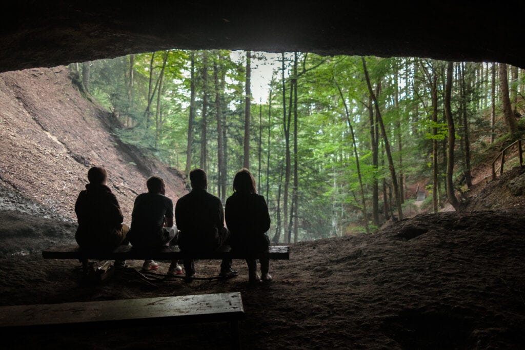 People sitting on a bench at the entrance to a cave in the woods.