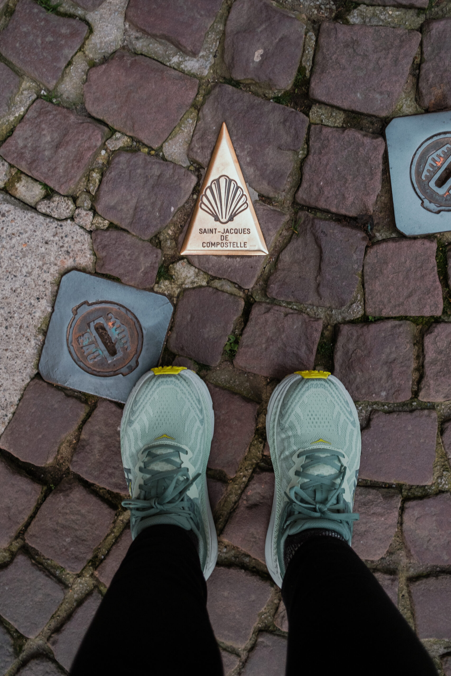 My feet beside a plaque on the road pointing the way on the Saint-Jacques de Compostelle.