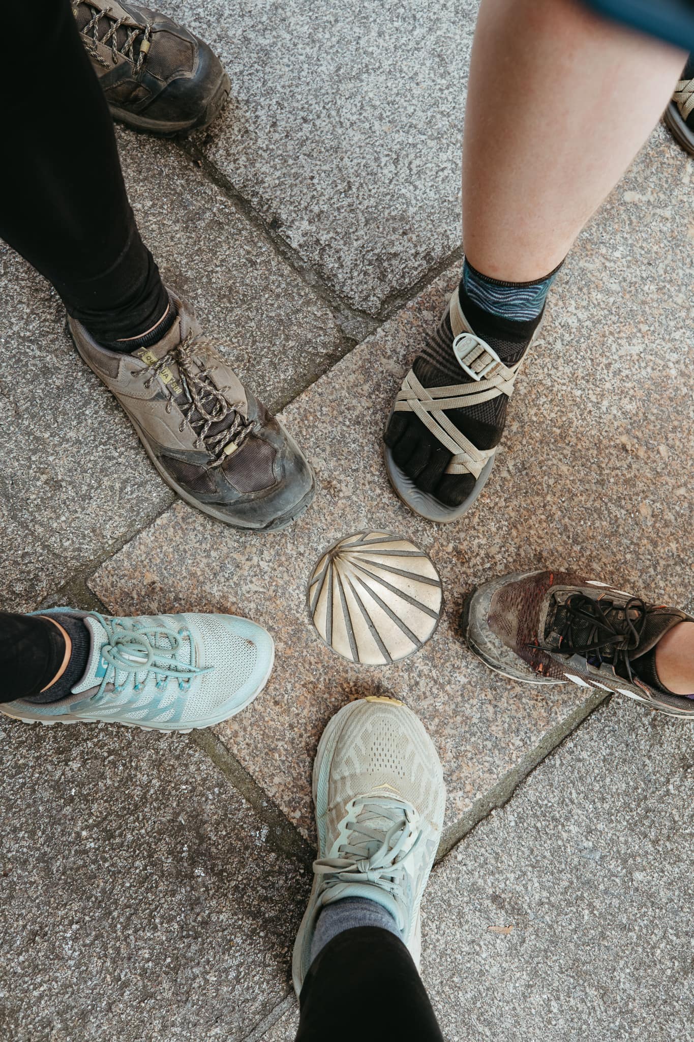 Five pilgrims' feet around a Camino shell in the ground.