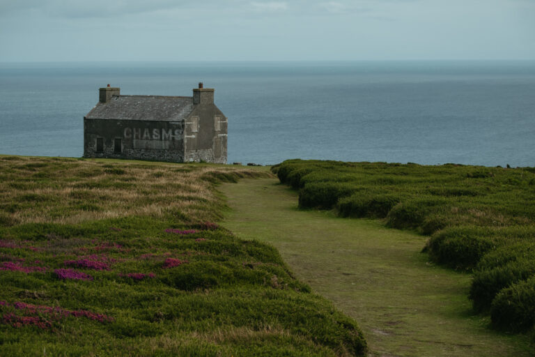 An abandoned gray building that says "Chasms" in the midst of green fields and in front of the ocean.