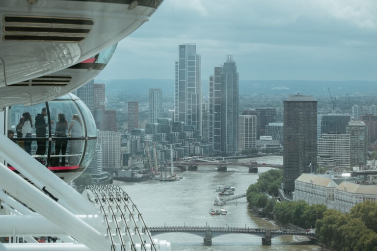 On the left side of the photo, people stand in a glass capsule. They are riding the London Eye. The remainder of the photo is filled with a river -- the Thames -- bridges across the river, and tall city buildings on either side of the river.