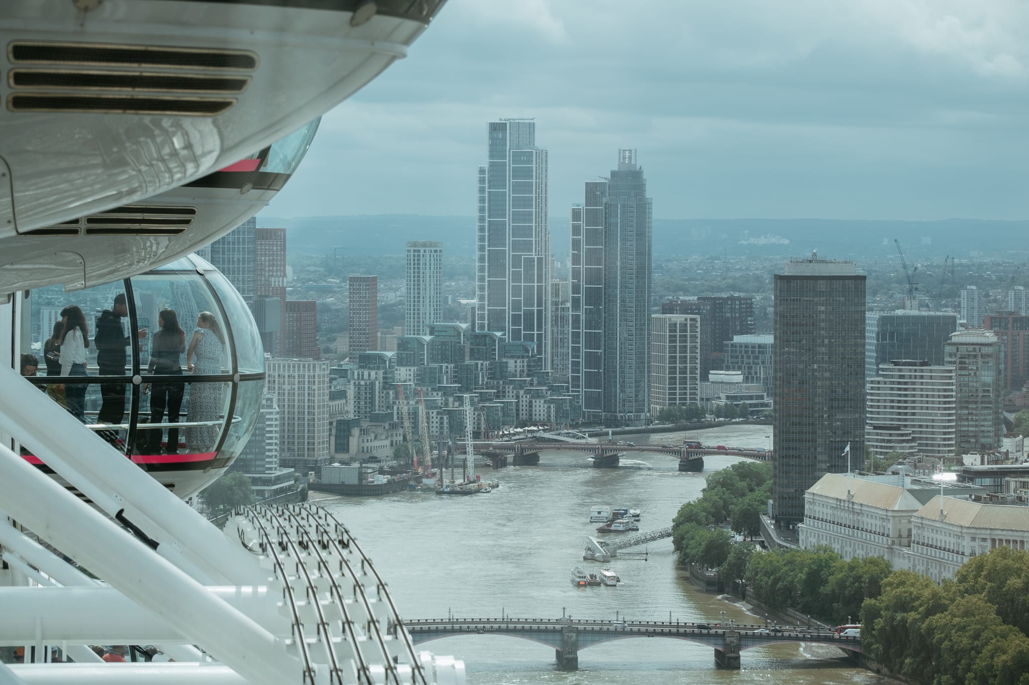 On the left side of the photo, people stand in a glass capsule. They are riding the London Eye. The remainder of the photo is filled with a river -- the Thames -- bridges across the river, and tall city buildings on either side of the river.