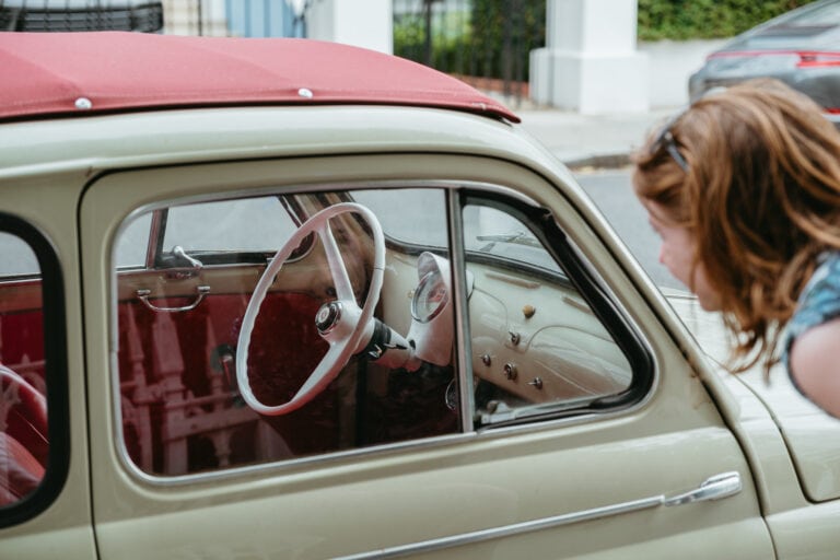 A red-haired woman peers through the window of a vintage car parked on the street in Notting Hill. The car is a sort of khaki color with red material on the roof. The steering wheel is white, and the interior of the car is the same khaki and red.