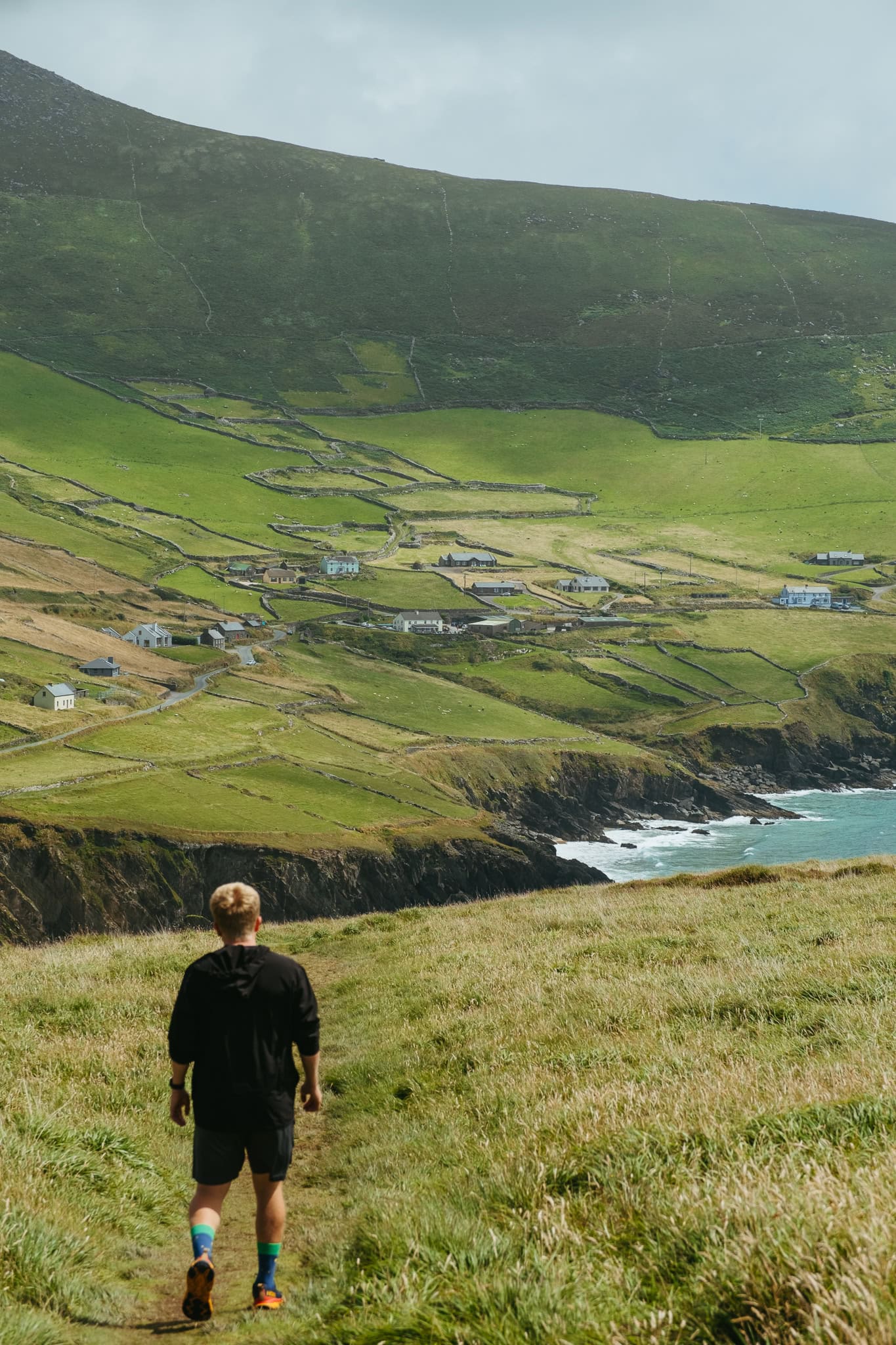 My brother walks down the hill at Dunmore Head, looking toward buildings on green hills above and turquoise water below.