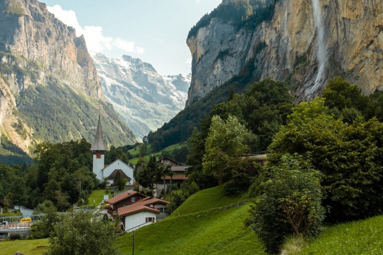 The picturesque little church of Lauterbrunnen sits amongst green hills and trees, beside a cliff with a huge waterfall.