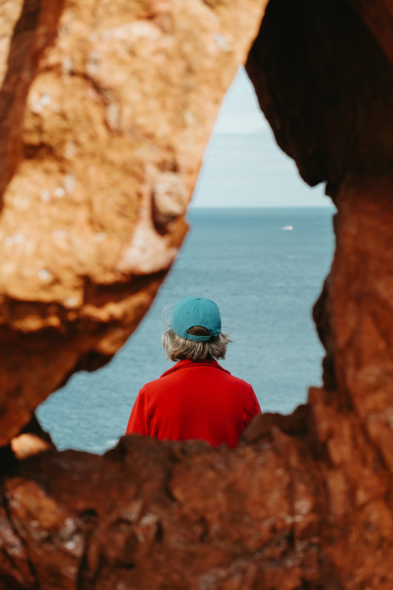 A woman in red, with a blue hat, stands looking out toward the sea.