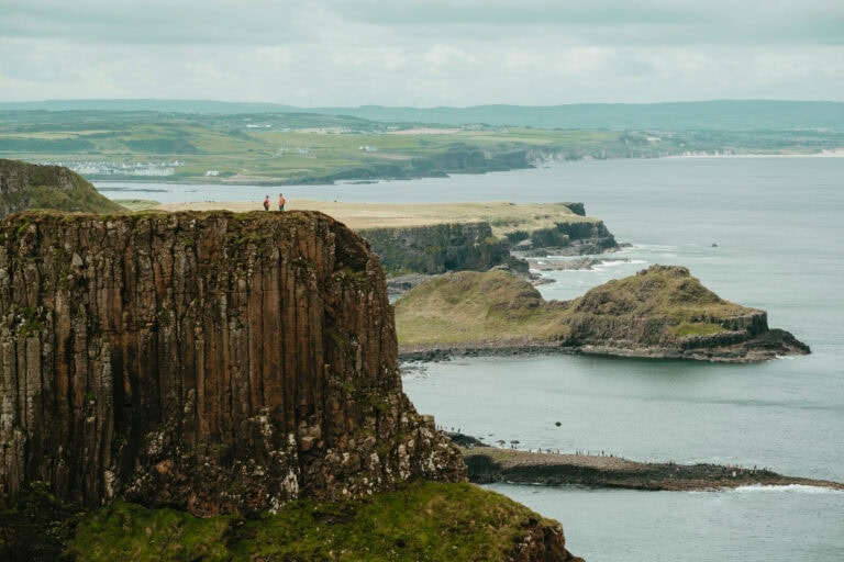 Two people stand on top a cliff above Giant's Causeway. In the distance is Northern Ireland's coastline.