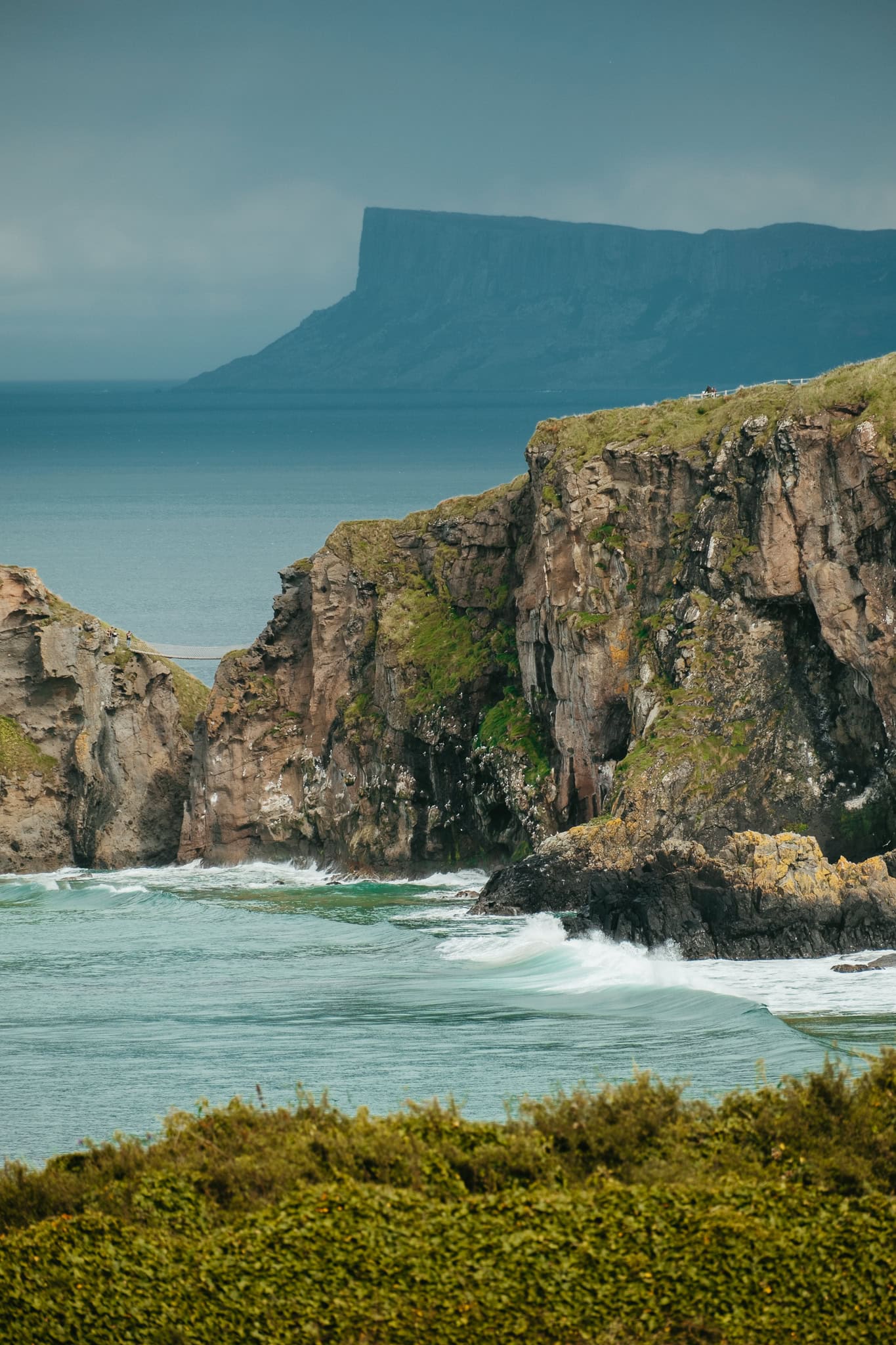Carrick-a-Rede Bridge, as seen from a distance. A sharp cliff is visible in the distance.