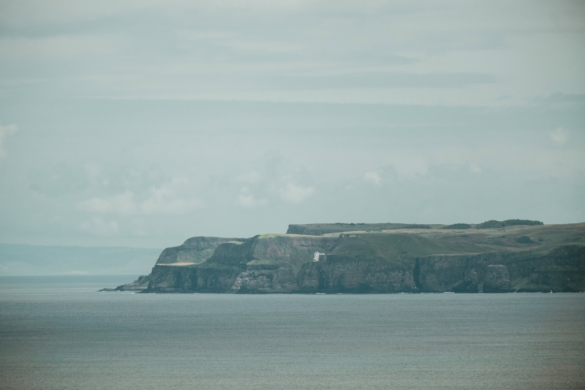 An island is distantly visible in the water. At the end of it, a white lighthouse sits on the cliff.