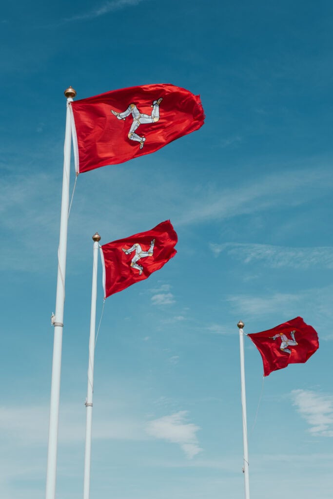 Three red Isle of Man flags flying in front of a blue sky.