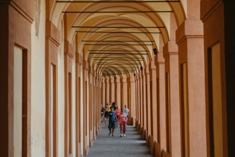 People walk through warm pink pillars of the Portico of San Luca.