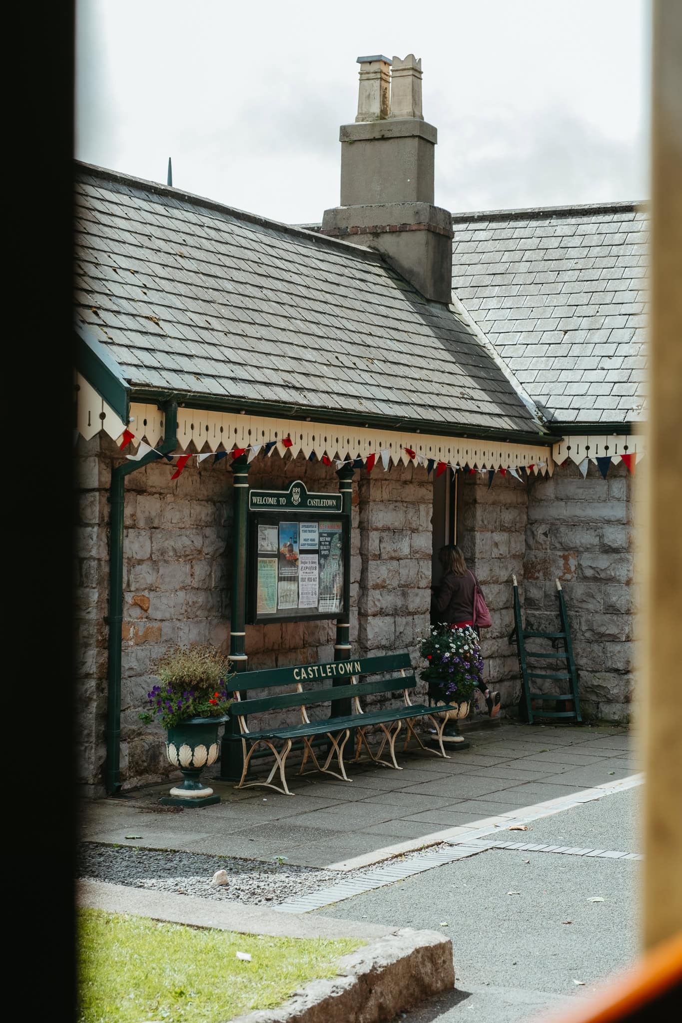 Through a train window, the small stone Castletown station is visible. There's a green bench that says "Castletown," colorful flags along the roofline, and several plants.