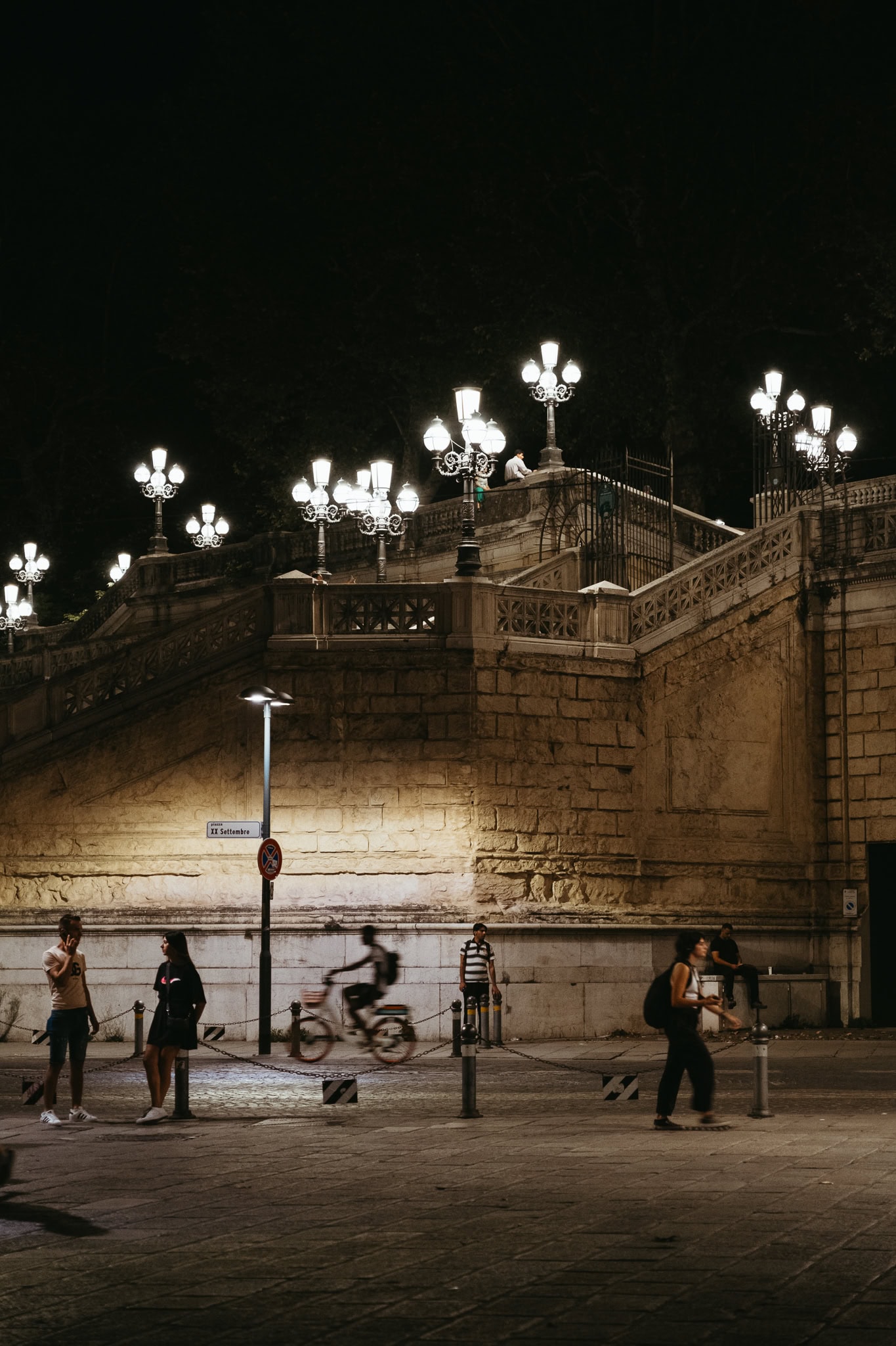 People walk and bike through a square in Bologna at night.