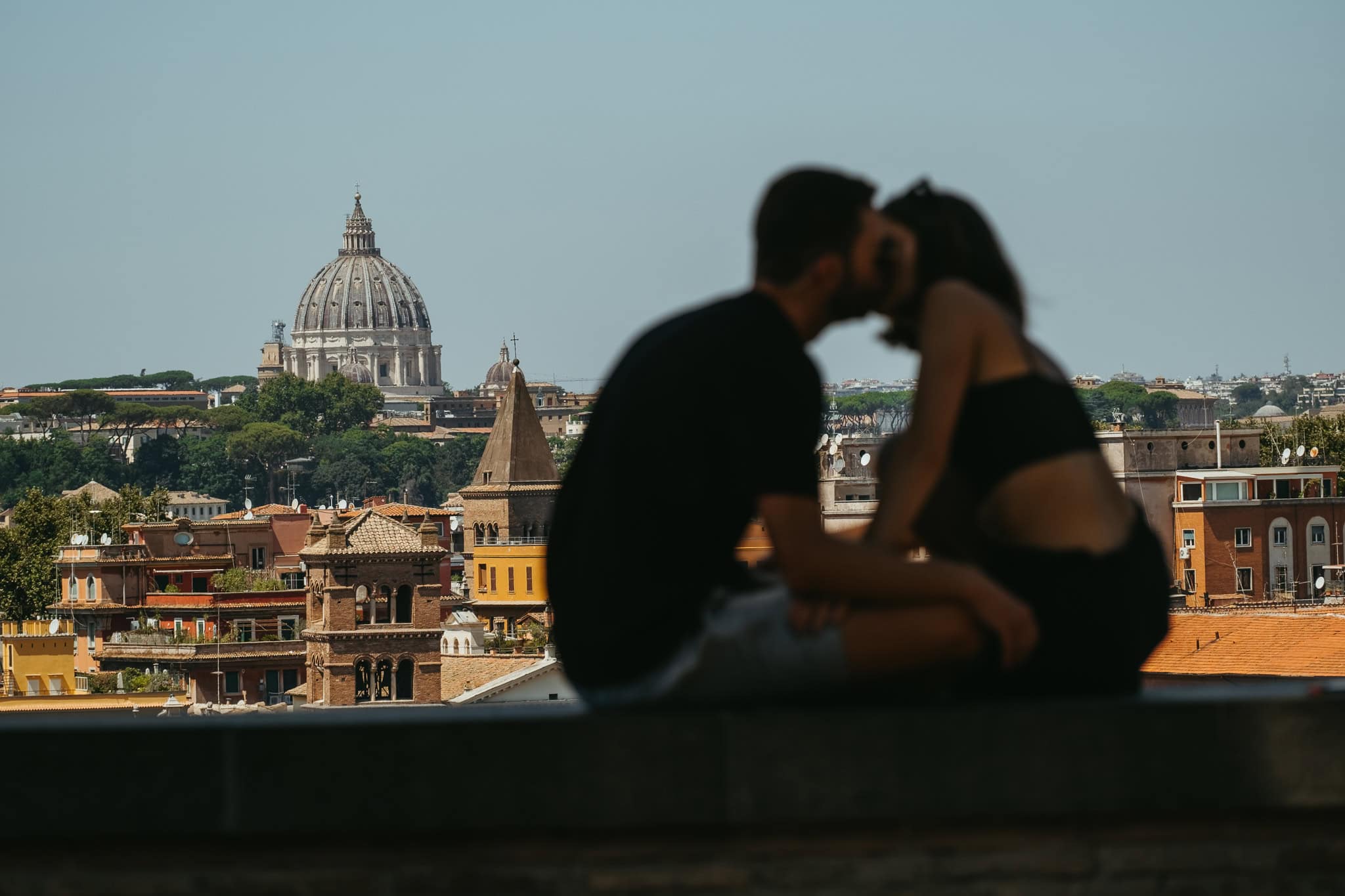 A couple kissing on top of a wall, with St. Peter's Basilica in the distance behind them.