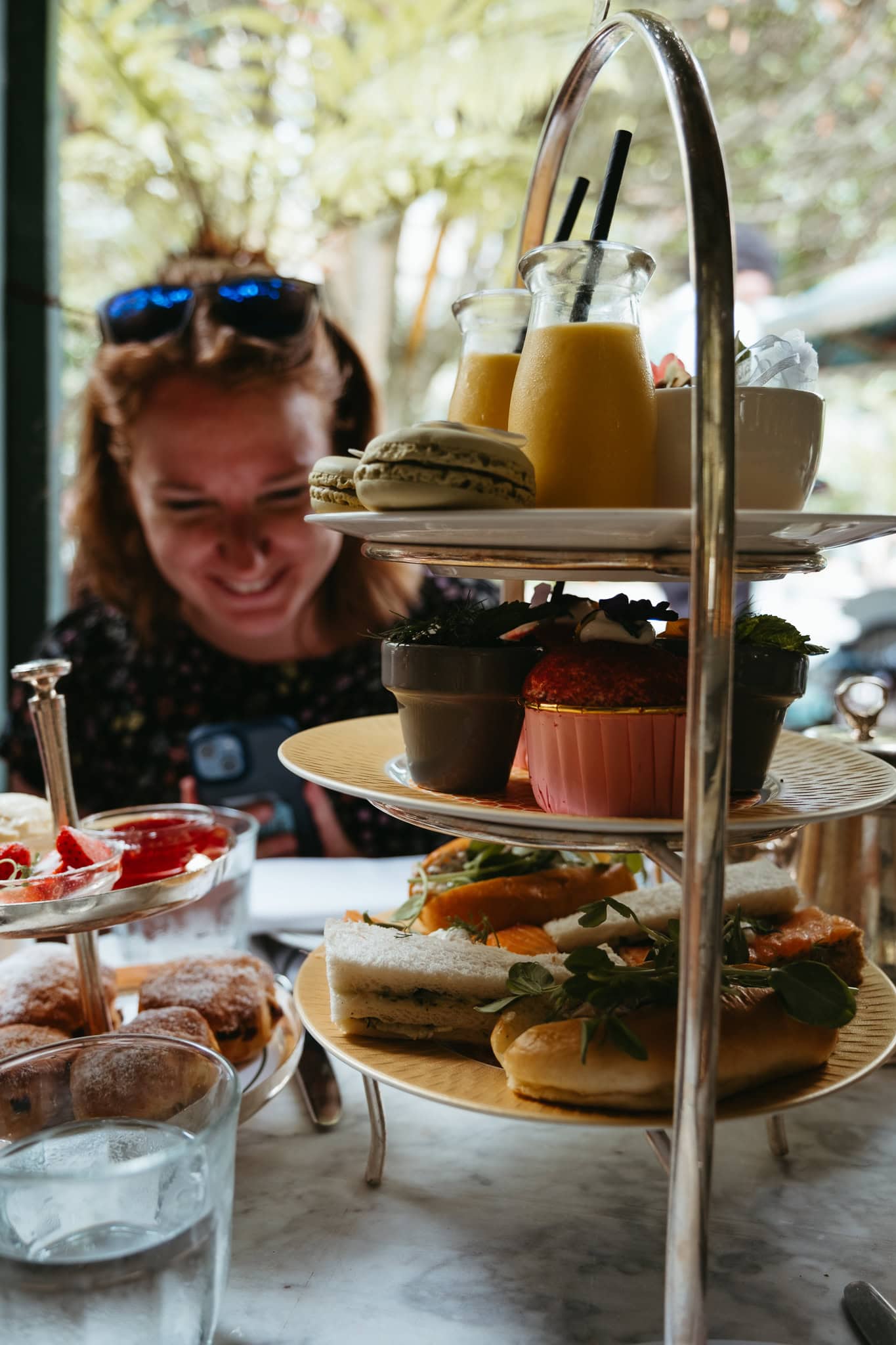 A woman smiles behind a tower of plates holding sandwiches, sweets, and more.