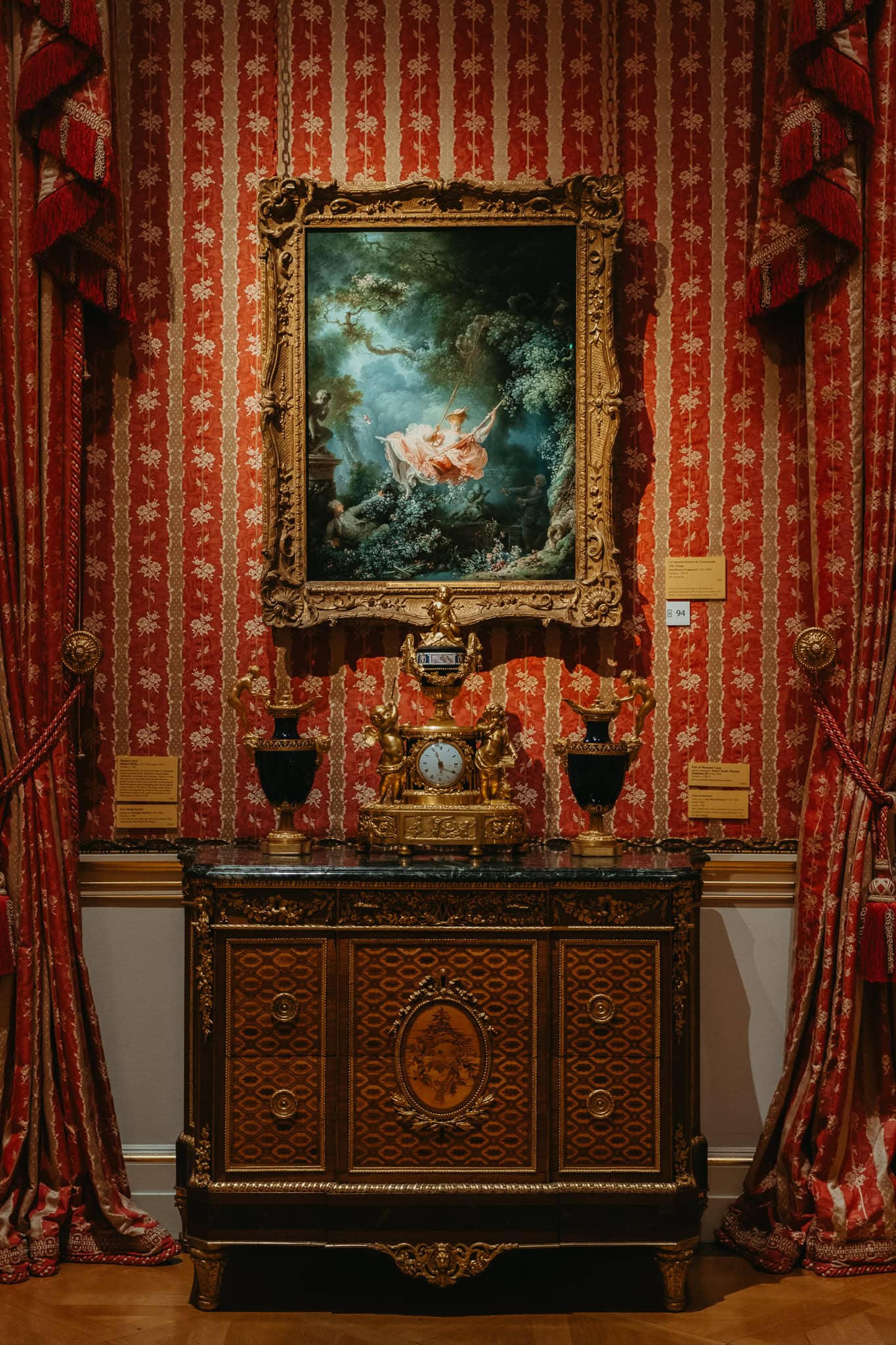 A painting of a young woman on a swing hangs on a red wall.