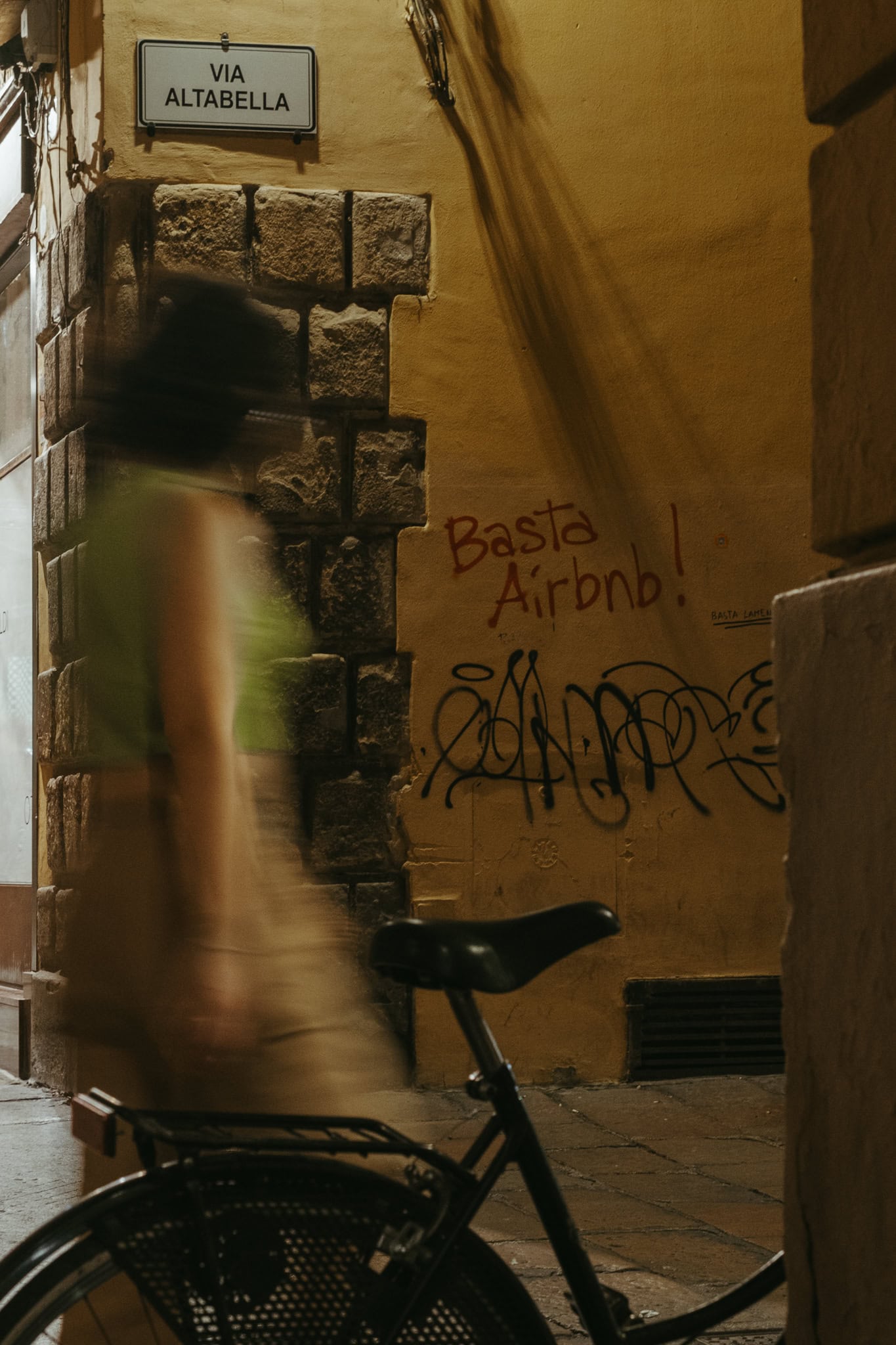 A blurry woman walks past a bicycle at night. On a wall behind her is written "basta Airbnb"