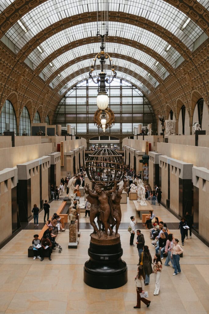 A long hall with an arched ceiling filled with statues on the ground level as people walk around.