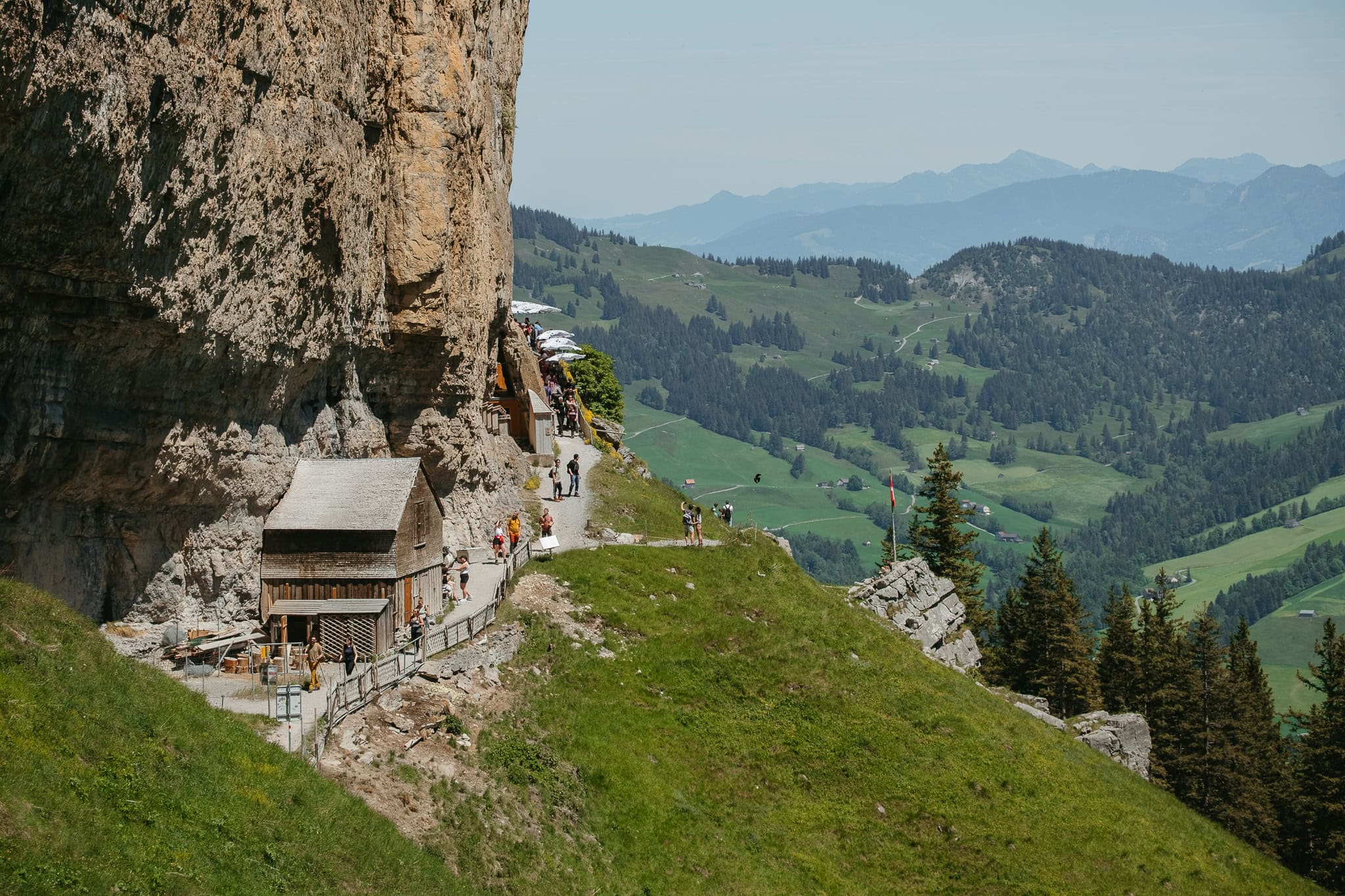 A small building at the side of a cliff and people walking along hiking trails.