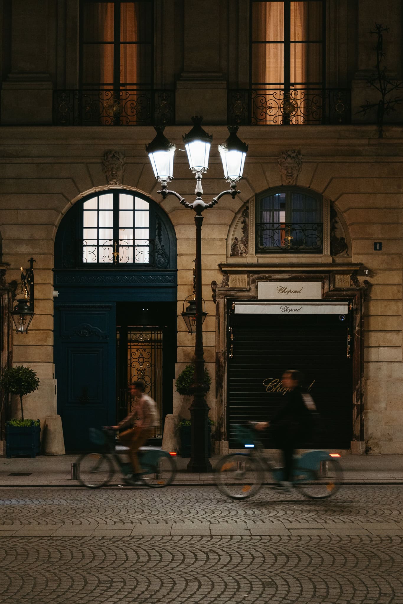 Two blurry bicyclists passing large rounded doors at night.