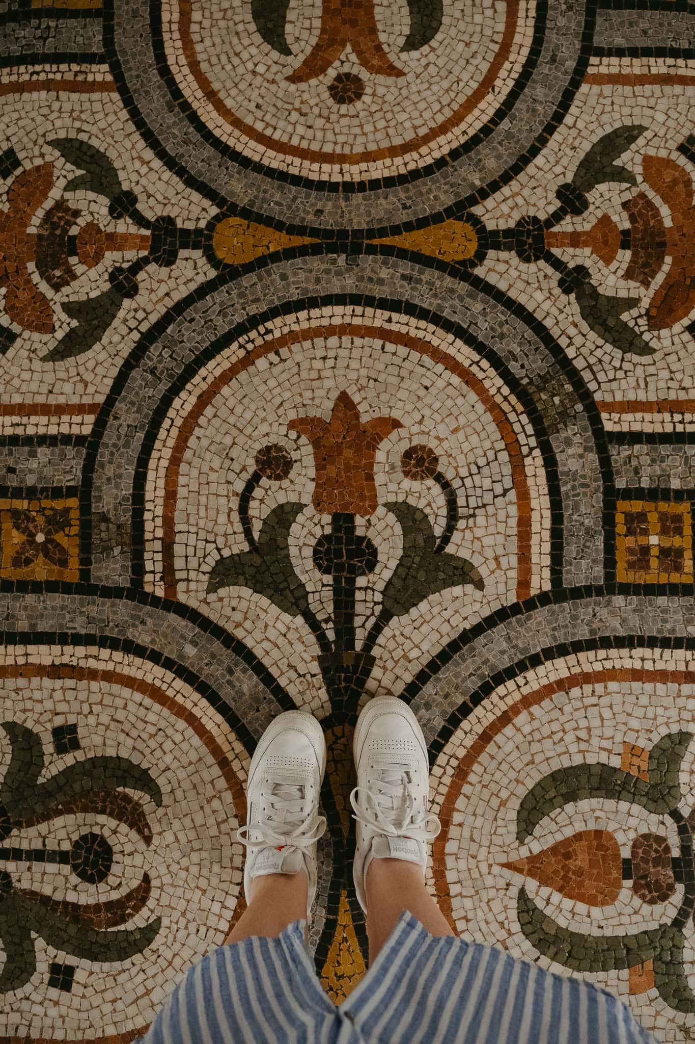 Feet in white sneakers standing on an ornate mosaic floor with flower designs.