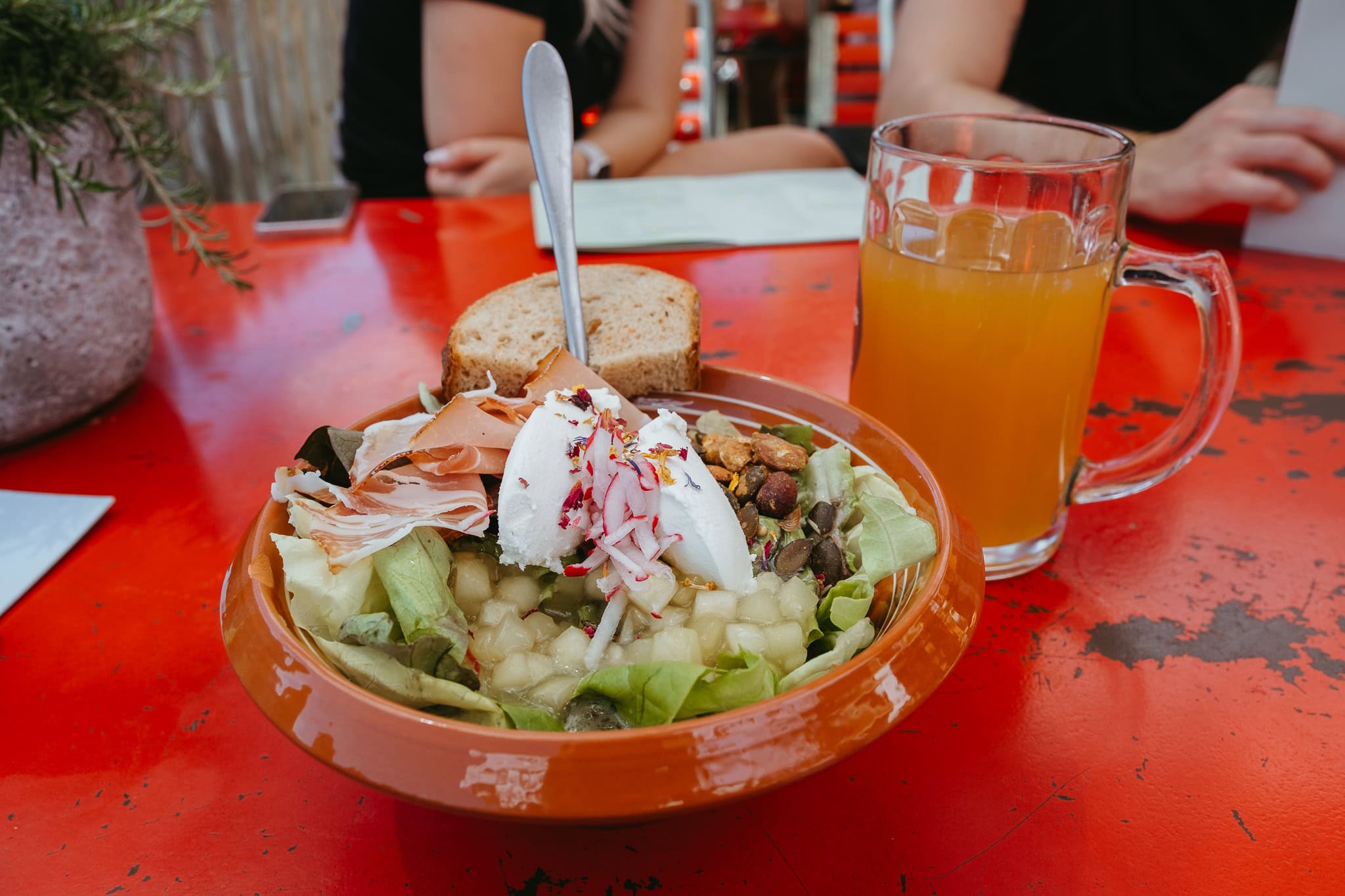 A salad with apple, sliced meats, nuts, cheese, and more sitting on a red table beside a glass of cider.