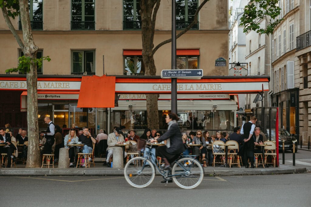 Woman rides bike in front of busy cafe in Paris.