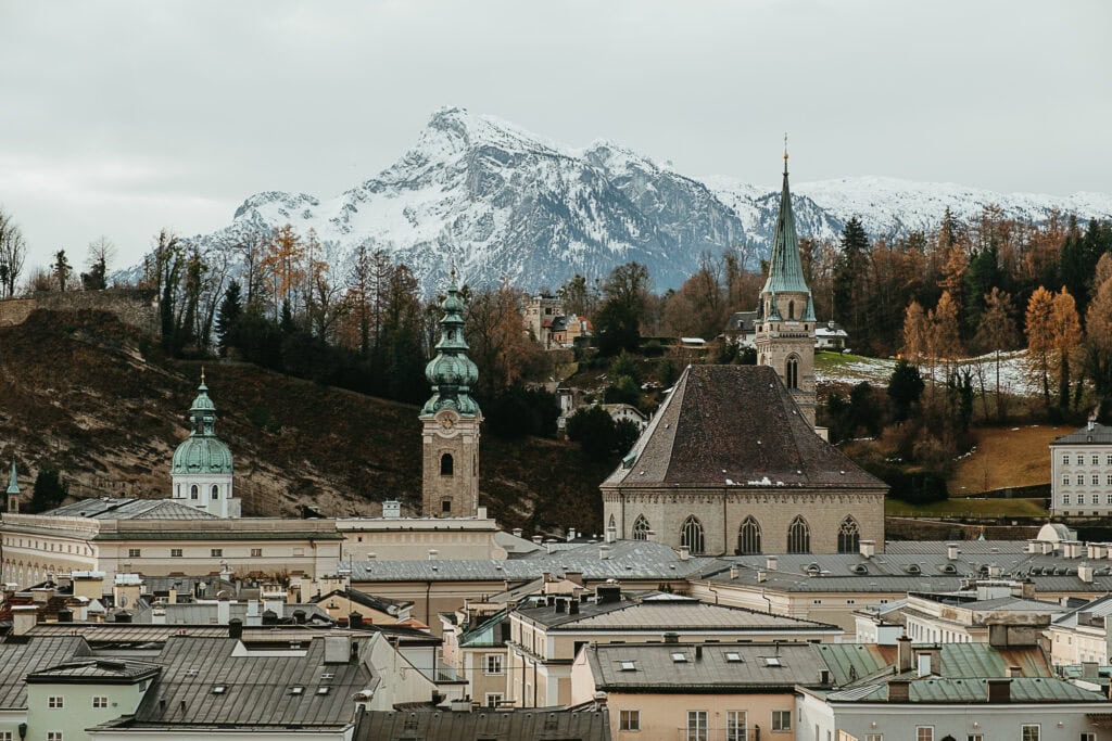 Church towers rising out of the rooftops of Salzburg with a snow-covered mountain in the background.